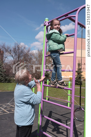 Smiling girl climbing colorful jungle gym while elderly woman watches and supports her from below Smiling girl climbing colorful jungle gym while elderly woman watches and supports her from below 125835947