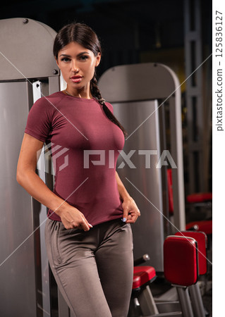 fitness enthusiast stands confidently in gym showcasing athletic wear while preparing begin intense workout session. Bright gym equipment surrounds her. 125836127