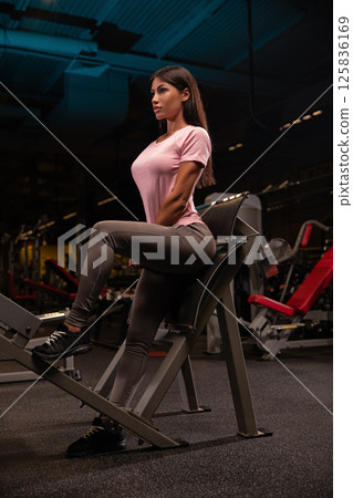 In modern gym setting young woman works out on leg extension machine emphasizing her fitness routine. gym features various exercise equipment and ambient lighting. 125836169