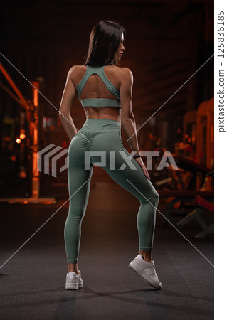 fitness model demonstrates athletic wear while posing confidently in gym setting during evening workout. atmosphere is energetic dim lighting highlighting her dedication. 125836185
