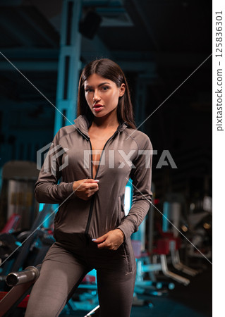 model stands confidently in stylish fitness outfit while posing in well equipped gym. setting features various workout equipment and prominent lighting. model stands confidently in stylish fitness outfit while posing in well equipped gym. setting features various workout equipment and prominent lighting. 125836301