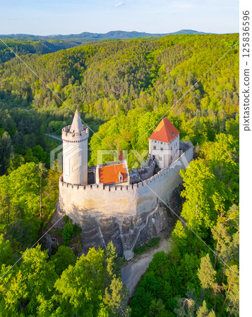 Kokorin Castle stands majestically amid lush green trees during a sunny evening. The aerial view captures the castle's distinct architecture and enchanting landscape. Kokorin Castle stands majestically amid lush green trees during a sunny evening. The aerial view captures the castle's distinct architecture and enchanting landscape. 125836596