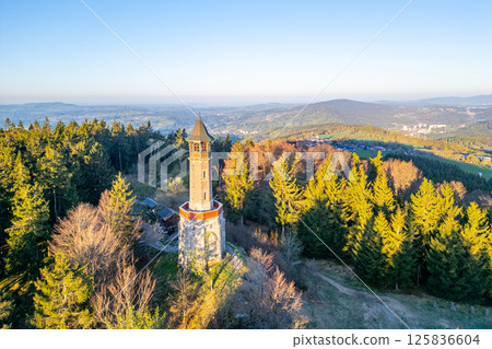 In a serene spring morning, the Stepanka Lookout Tower stands tall amidst lush green trees in Korenov, offering expansive views of the Jizera Mountains and surrounding landscape. 125836604