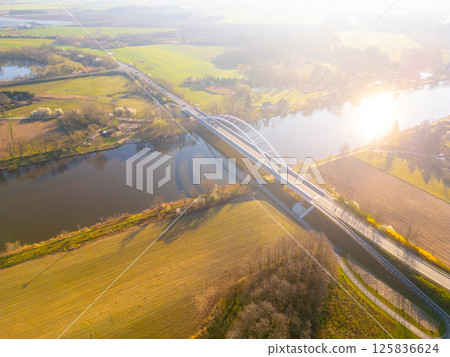Bohumil Hrabal's Bridge gracefully stretches over the Labe River in Litol, Lysa nad Labem, Czechia, surrounded by lush greenery and an expansive landscape during golden hour. 125836624
