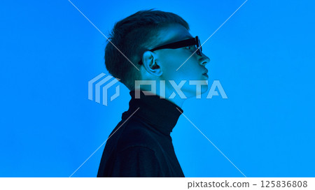 Side profile of stylish young man in turtleneck and sunglasses, posing under deep neon blue background ,expresses futuristic elegance. 125836808