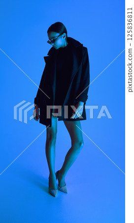 Full-length portrait of confident young woman in coat, heels and sunglasses posing against blue background in neon light. 125836811