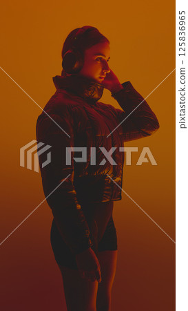 Side view portrait of young woman listening to music in headphones, wearing puffer jacket against orange background in neon lights. 125836965