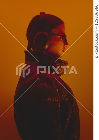 Woman wearing headphones, black jacket and sunglasses, posing against orange studio background in neon. Contemporary style and music. 125836966