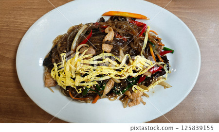 Delicious Plate of Korean Japchae with Stir-Fried Glass Noodles, Vegetables, meat, and Egg Garnish on a White Plate, Captured on a Wooden Table in a Cozy Restaurant 125839155