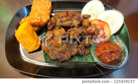 Spicy Grilled Oxtail with Tofu and Tempeh Served with Fresh Cucumber and Tomato Slices and Spicy Chili Sauce on Black Plate 125839237