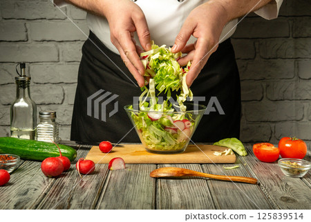 Hands of a chef mix fresh vegetables including lettuce, radishes, and cucumbers in a glass bowl on a wooden cutting board Hands of a chef mix fresh vegetables including lettuce, radishes, and cucumbers in a glass bowl on a wooden cutting board 125839514