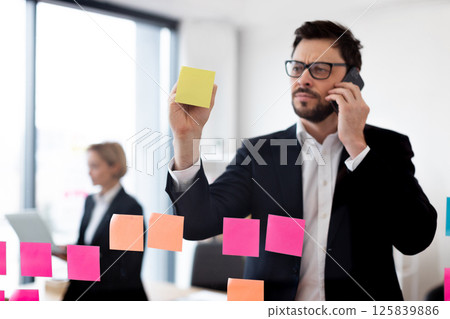 Young adult male professional in business suit uses colorful sticky notes on glass panel while talking on mobile phone in modern office environment emphasizing organization 125839886