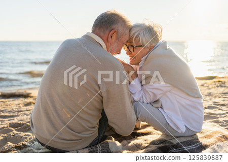 Senior mature couple sitting on beach holding hands enjoying outdoor recreation. Old husband wife touching hands with tenderness love. Grandmother grandfather together. Family moment of love 125839887