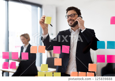 Businessman wearing glasses writing on sticky notes on transparent board while on phone. Professional setting suggesting planning organization and productivity. Female colleague in background working. 125839888