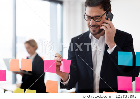 Young adult male using colorful sticky notes for business task organization on glass board. Holding phone engaged in conversation in modern office setting. Young adult male using colorful sticky notes for business task organization on glass board. Holding phone engaged in conversation in modern office setting. 125839892