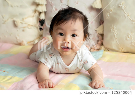 Adorable Asian Baby in Easter Themed Onesie Lying on Pastel Blanket and Smiling on Bed Decorated with Embroidered Pillows and Soft Textures Warm Natural Light and Cozy Atmosphere 125840004