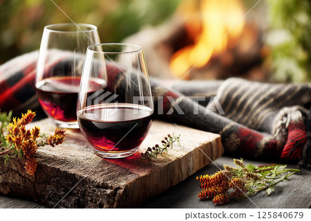 Warm evening with red wine glasses placed on a wooden board beside a cozy fire in nature embrace. 125840679