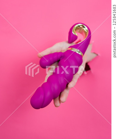 Female hand sticking out with pink sex toy from pink paper background. Vertical photo. Female hand sticking out with pink sex toy from pink paper background. Vertical photo. 125841683