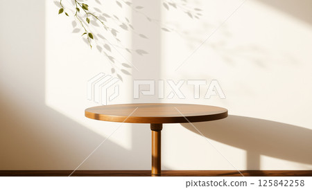A simple wooden table with a round top filled with a coating that provides sunlight. The delicate shadows of leafy branches play on the textured cream wall in the background. A simple wooden table with a round top filled with a coating that provides sunlight. The delicate shadows of leafy branches play on the textured cream wall in the background. 125842258