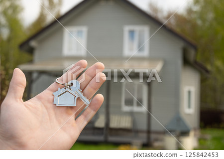 Man hand holds house-shaped key in front of newly purchased home. New beginning, homeownership, and real estate acquisition, representing success and investment. Real estate agent 125842453