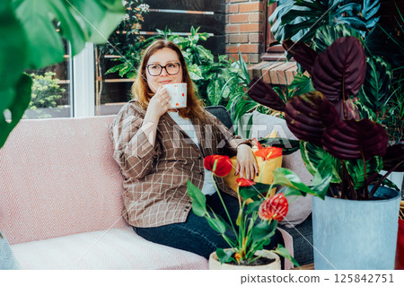 Smiling woman with cup of drink relaxing on sofa in home garden conservatory after housekeeping. Portrait of relaxed female resting at home. Gardening hobby. Urban jungles. Biophilia lifestyle. Smiling woman with cup of drink relaxing on sofa in home garden conservatory after housekeeping. Portrait of relaxed female resting at home. Gardening hobby. Urban jungles. Biophilia lifestyle. 125842751