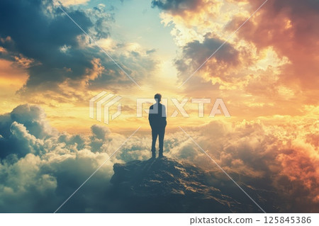 A man stands on a mountain top, looking out at the sky and the sun is setting in the distance. Scene is peaceful and contemplative 125845386
