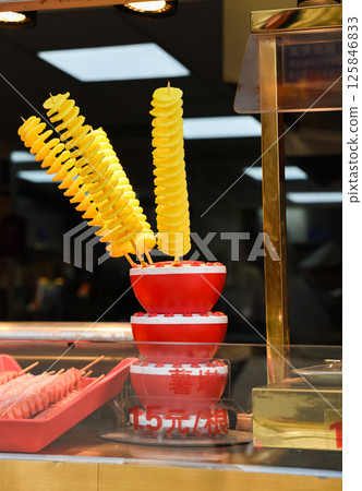 A popular Chinese street snack in Shanghai - golden spiral-cut tornado potatoes on wooden skewers, displayed in stacked red bowls at a vibrant market stall. 125846833