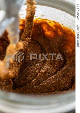 The mixer bowl shows a rich, dark molasses added to the creamed butter and sugar. The molasses is beginning to integrate, creating a deep brown, aromatic mixture. The mixer bowl shows a rich, dark molasses added to the creamed butter and sugar. The molasses is beginning to integrate, creating a deep brown, aromatic mixture. 125847153
