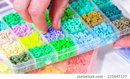 Womans hands gracefully poised over a collection of beads, sorted by color in transparent organizers. The array of beads spans a vibrant spectrum, from deep purples to bright oranges, meticulously 125847177