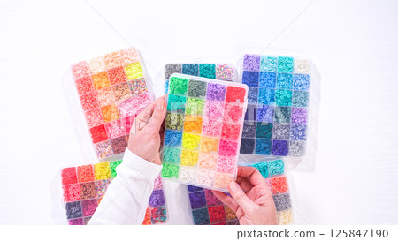 Flat lay. Womans hands gracefully poised over a collection of beads, sorted by color in transparent organizers. The array of beads spans a vibrant spectrum, from deep purples to bright oranges 125847190