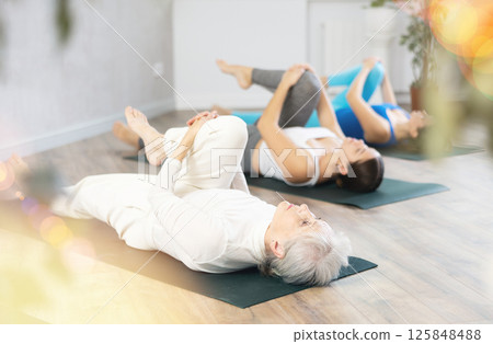 Active old woman stretching gluteal muscle during group yoga Pilates class on mats in gym studio Active old woman stretching gluteal muscle during group yoga Pilates class on mats in gym studio 125848488