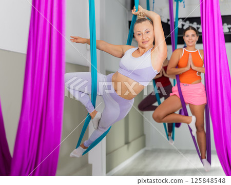 Young woman hanging upside down while practicing aerial yoga during group class in modern fitness club 125848548
