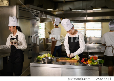 Busy cooking class in session with multiple students in uniforms preparing fresh ingredients Busy cooking class in session with multiple students in uniforms preparing fresh ingredients 125848641