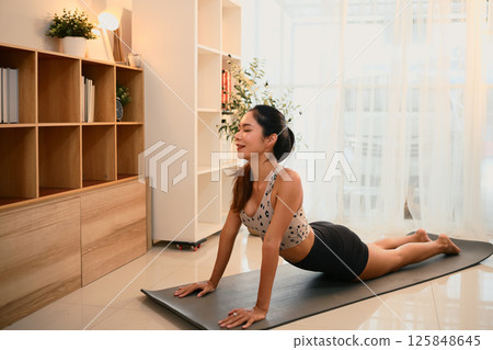 Fit woman doing cobra pose on exercise mat during a home yoga session Fit woman doing cobra pose on exercise mat during a home yoga session 125848645