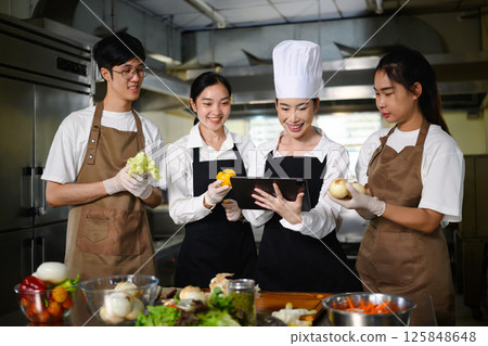 Smiling chef teacher using a tablet to explain a recipe to students in a professional kitchen 125848648