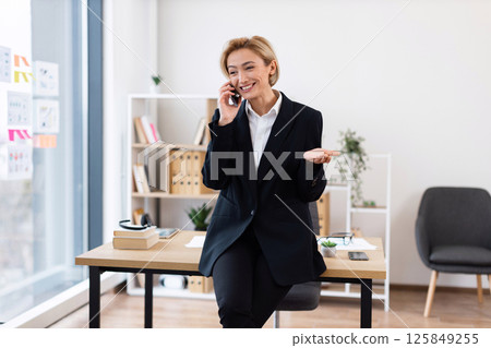Confident woman in her 30s, dressed in formal attire, chatting on phone in stylish office setting with desk and shelves in background, conveying business success and communication 125849255