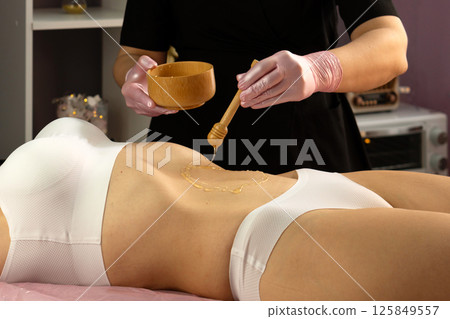 Beautician applies honey to abdomen with dipper during spa session. 125849557