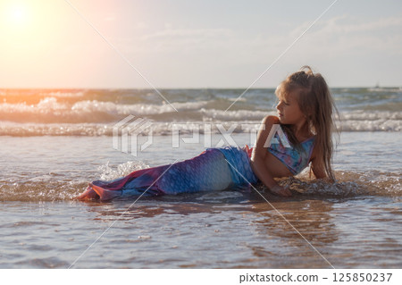 A girl in a mermaid costume on the beach. Sunset on the shore of the blue sea. Childhood happiness, beach, vacation, sunscreen, warm weather, travel. High quality photo 125850237