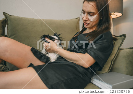 woman in black robe with under eye patches gently petting sleepy white black japanese chin dog on green sofa in cozy home lamp and houseplant, wellness, pets care, lifestyle brands 125850756