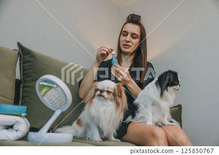 woman applying facial serum with small dropper, morning beauty routine, sitting on green sofa with two japanese chin dogs on her legs 125850767