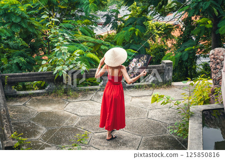 back of beautiful woman tourist traveler in Vietnamese non la hat with a fan in a garden in Vietnam. Tourism and travel in Asia 125850816