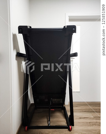 Folded black treadmill positioned against a wall in a modern interior space with wooden flooring and door Folded black treadmill positioned against a wall in a modern interior space with wooden flooring and door 125851909