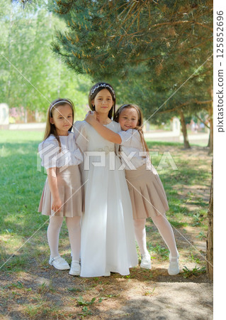 A little girl in a long ivory dress stands on a meadow among coniferous trees. Two twin girls in festive outfits gently hug the older sister 125852696