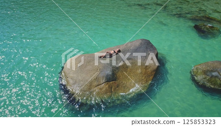 A person is lounging on a large rock in the middle of clear turquoise water. The sun shines brightly, highlighting the vibrant colors of the scene and creating a tranquil atmosphere. A person is lounging on a large rock in the middle of clear turquoise water. The sun shines brightly, highlighting the vibrant colors of the scene and creating a tranquil atmosphere. 125853323