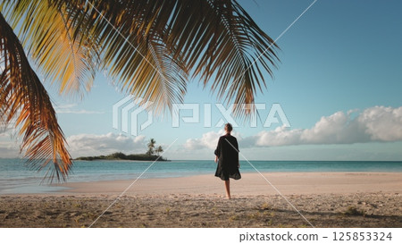 Young woman is enjoying a peaceful moment on a tropical beach, with palm trees swaying gently in the background and the sun setting over the ocean 125853324