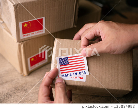Man Hands applying MADE IN USA over MADE IN CHINA label on a shipping cardboard box. 125854998