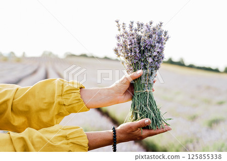 woman in yellow dress stands in lavender field, gently holding bouquet of lavender flowers. She appears serene and contemplative, enjoying beauty and fragrance of blossoms around her. 125855338