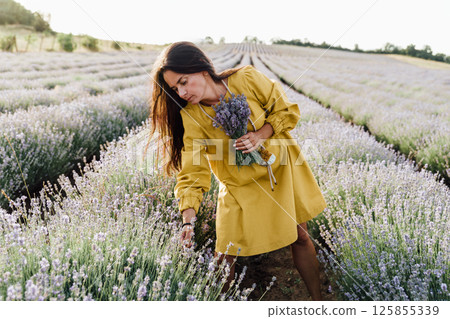 woman in yellow dress stands in lavender field, gently holding bouquet of lavender flowers. She appears serene and contemplative, enjoying beauty and fragrance of blossoms around her. 125855339