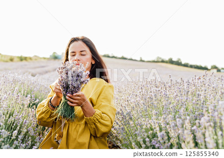 woman in yellow dress stands in lavender field, gently holding bouquet of lavender flowers. She appears serene and contemplative, enjoying beauty and fragrance of blossoms around her. woman in yellow dress stands in lavender field, gently holding bouquet of lavender flowers. She appears serene and contemplative, enjoying beauty and fragrance of blossoms around her. 125855340