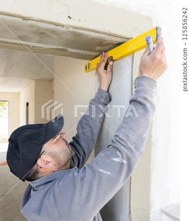Craftsman in grey work clothing uses a yellow level to control a house wall. Construction process Craftsman in grey work clothing uses a yellow level to control a house wall. Construction process 125856242
